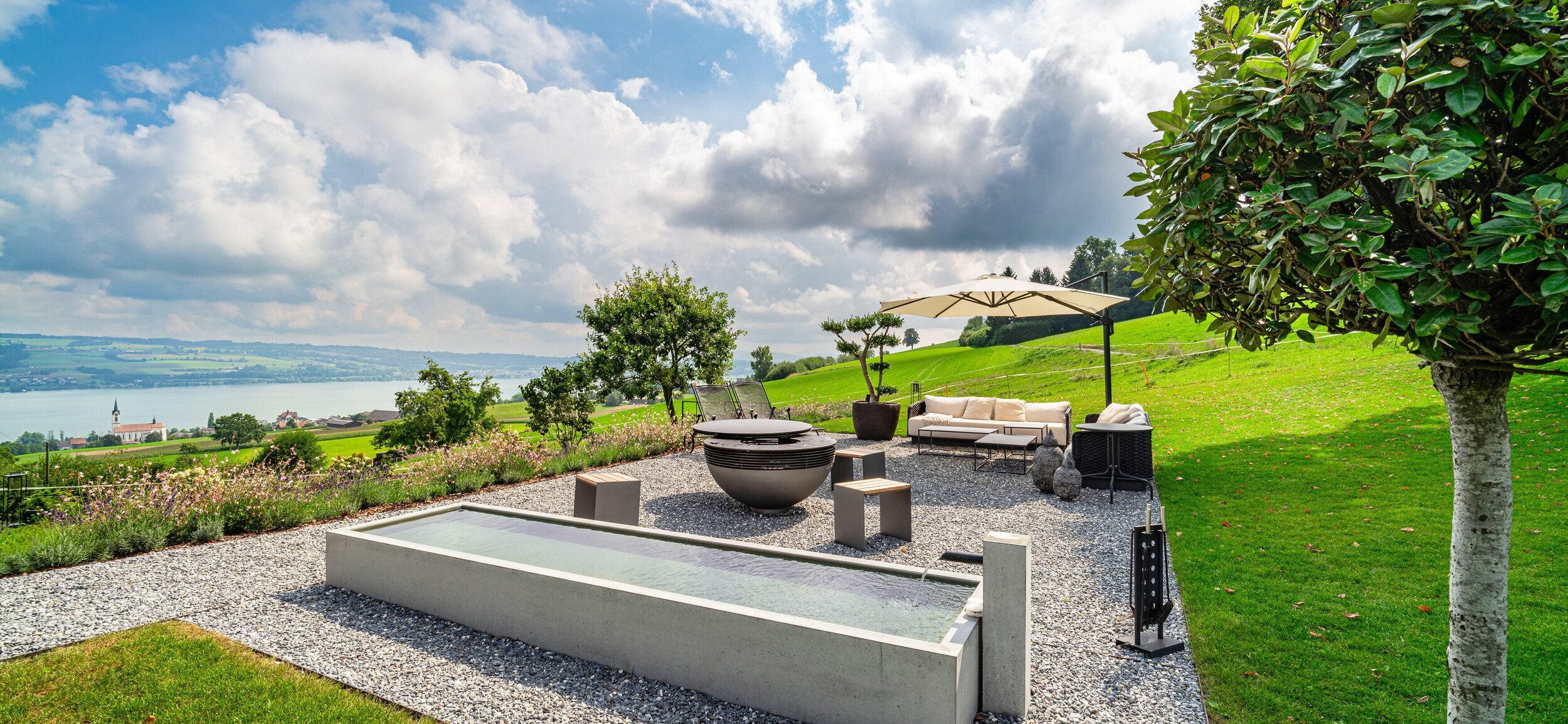 brunnen_aus_beton_von_creabeton_mit_kiesplatz_und_feuerschale_lounge_und_sonnenschirm