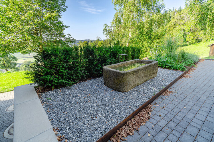 historischer_steinbrunnen_mit_kiesfläche_und_hecken_umgeben_von_naturreicher_landschaft_frieda_pflasterstein_crea_beton