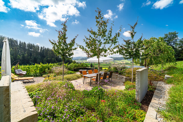 gartenbereich_mit_essenstisch_sandtsienplatten_schattenspender_und_blühender_bepflanzung_im_vordergrund_mit_grünem_landschaftshintergrund