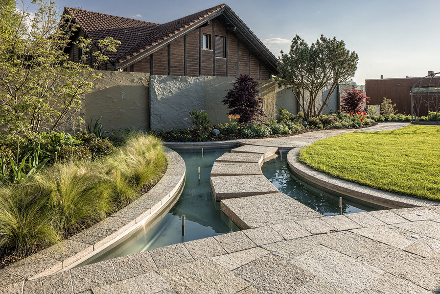 garten_mit_wasserlauf_und_steinen_im_modernen_design_umgeben_von_grünem_terrain_und_bäumen