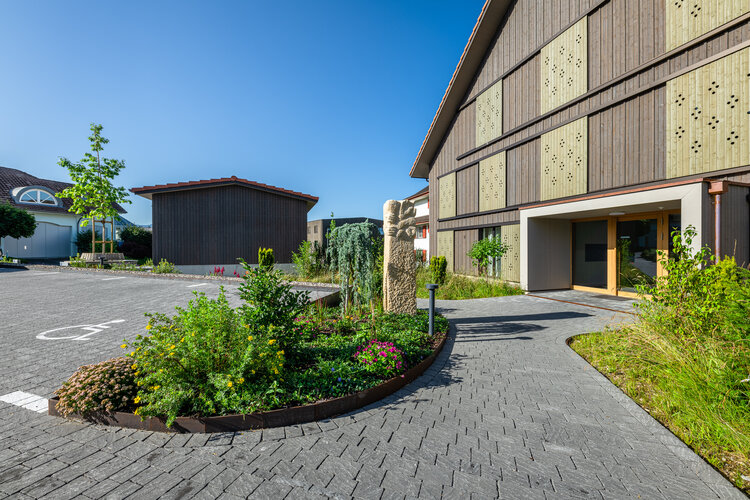 Eingangsbereich_eines_öffentlichen_Gebäudes_mit_modernem_Gartenbau_bepflanztes_Rundbeet_mit_Blumen_und_Sträuchern_gepflasterte_Gehwege_und_eine_markante_Steinskulptur_vor_einer_Holzfassade