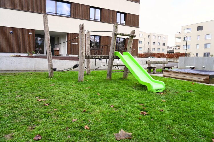spielplatz_mit_spielturm_und_sandkasten_im_rasen_mit_modernem_mehrfamilienhaus_im_hintergrund