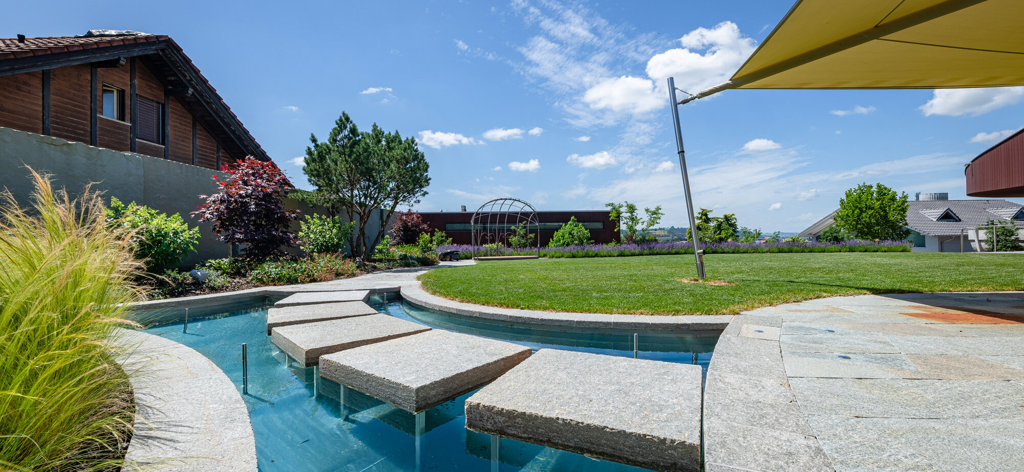 garten_mit_wasserbecken_und_steinplatten_aus_luserna_über_dem_wasser_umgeben_von_grünem_terrain_und_entspanntem_ausblick