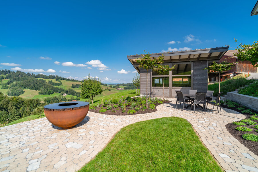 Gartenplanung_mit_Sitzbereich_und_Feuerstelle_in_malerischer_Landschaft_mit_grünen_Hügeln_und_blauem_Himmel