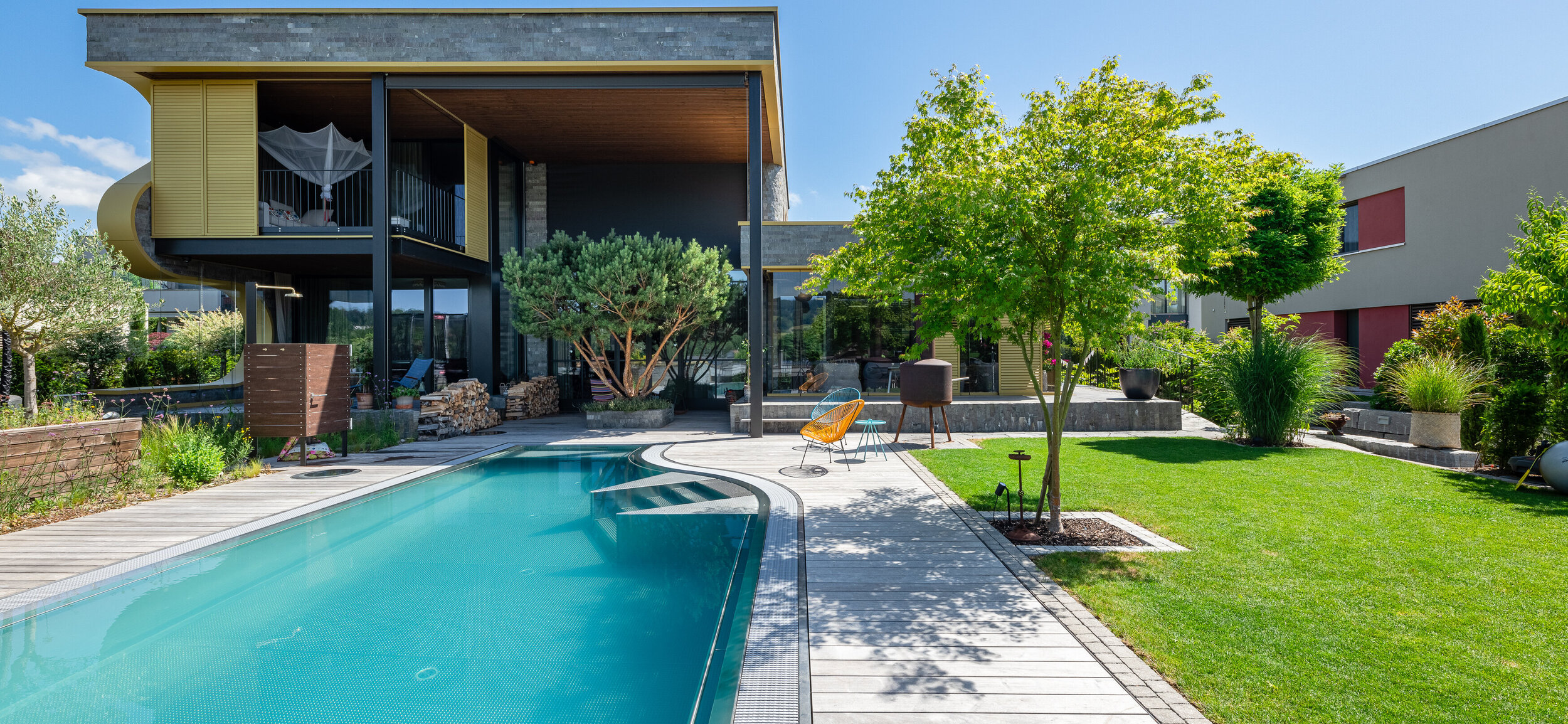 moderner_garten_mit_pool_terrasse_und_grüner_solitärstrauch_schattenbaum_rasenfläche_vor_architektonisch_modernem_haus