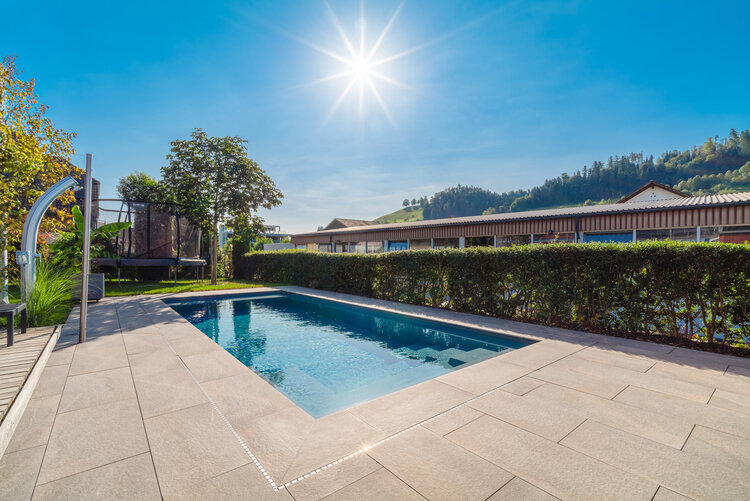 garten_mit_poolbereich_umgeben_von_hecken_und_grünem_terrain_im_hintergrund_moderne_gebäude_und_hügelige_landschaft
