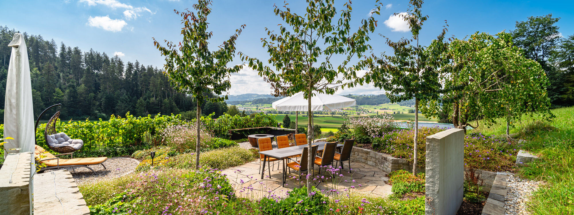 garten_mit_sandsteinkrustenplatten_grüner_bepflanzung_umgeben_von_blühenden_pflanzen_und_grünem_terrain